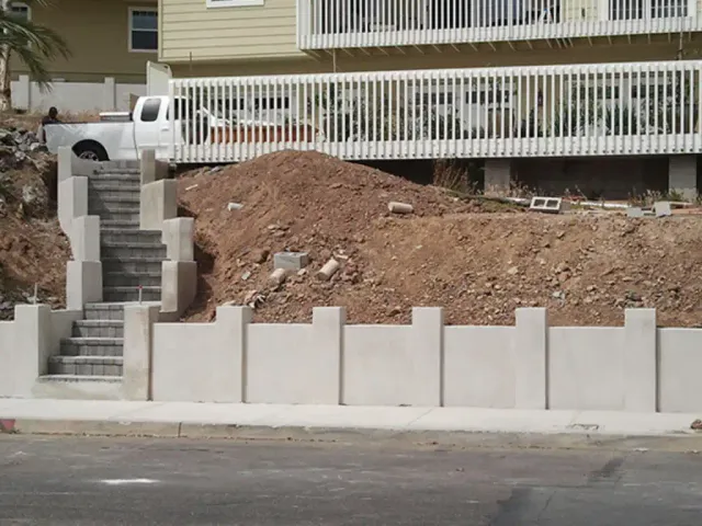 A finished stucco block wall with raised pilasters along a sidewalk, with a block stairway climbing the slope behind it.