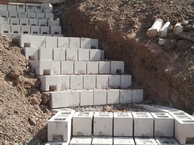 Concrete blocks being laid as stepped courses up a cut in reddish-brown soil.