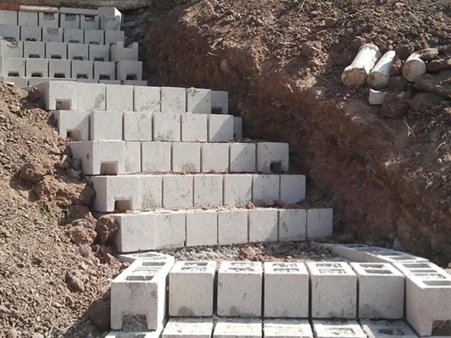 Excavated clay hillside with concrete block footing courses stepped into the cut and perforated drain pipe staged on the soil.