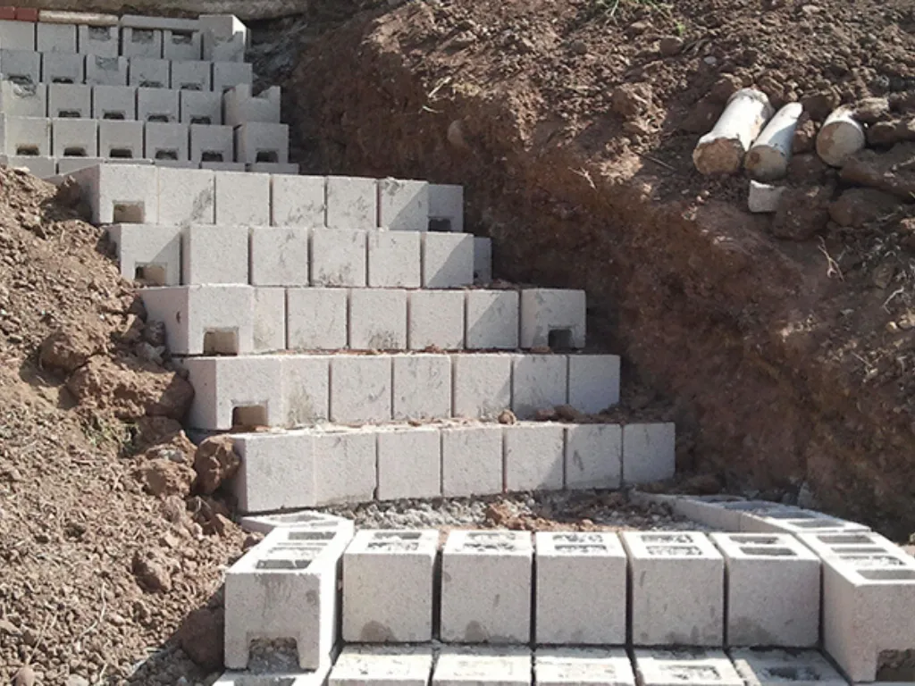 Stepped CMU block footing courses laid into an excavated hillside trench with perforated drain pipe staged alongside.