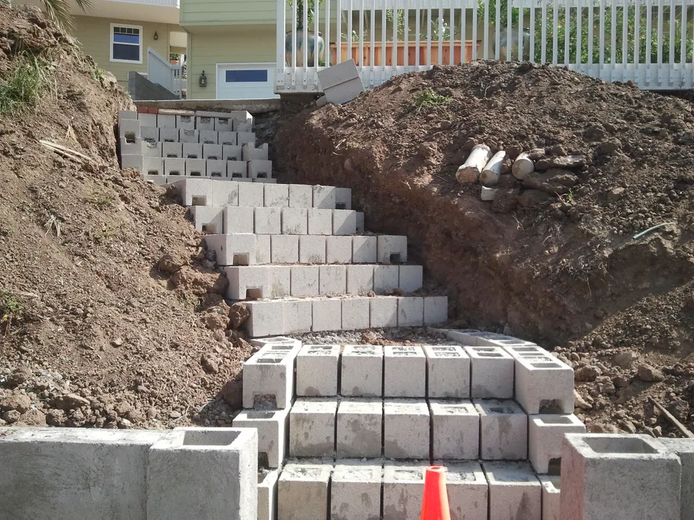 Stepped concrete block stair and walls under construction in a clay hillside cut, with drain pipe staged on the excavated soil.