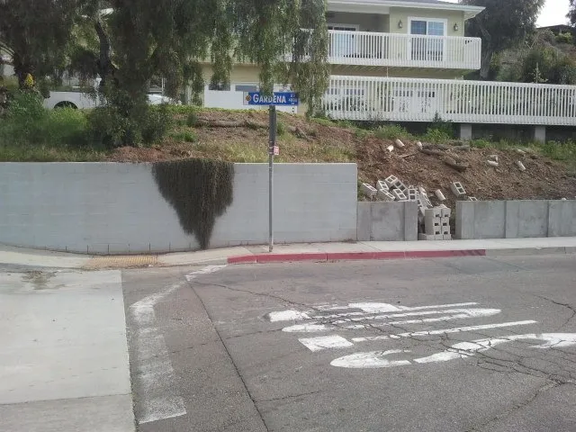 Old concrete block wall at a street corner below a sloped yard, with new block staged on the hillside.